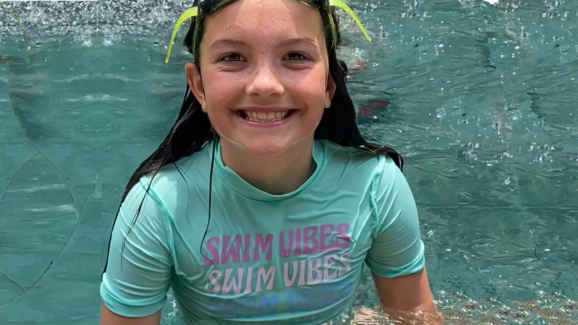 A smiling young swimmer in a swim shirt and goggles standing in a clear turquoise pool.