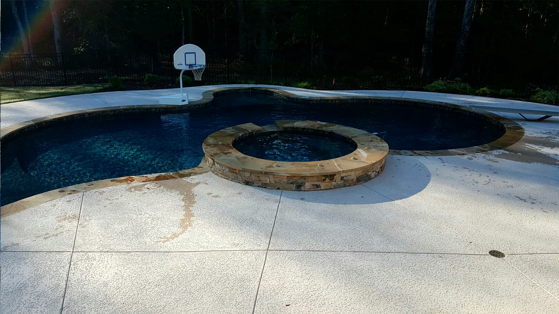 A freeform pool with a raised stone spa and a poolside basketball hoop, set in a broad concrete deck.
