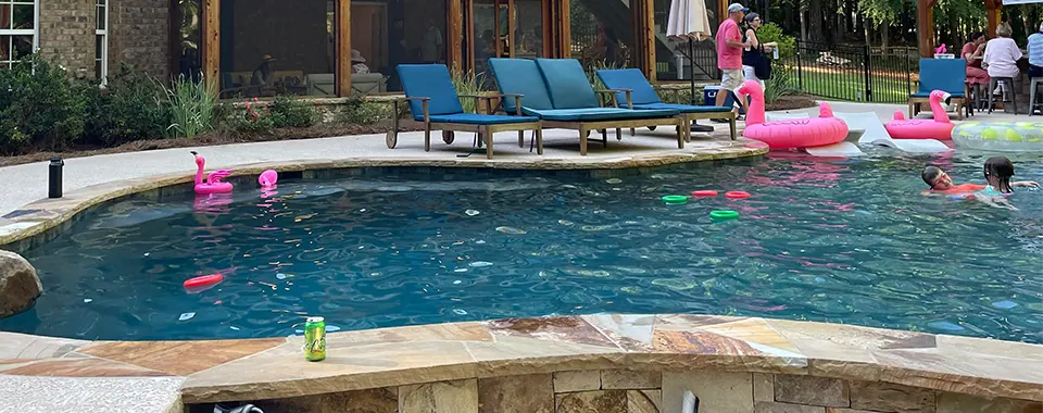 A backyard pool with pink flamingo floats, blue loungers on the deck and swimmers in the water.