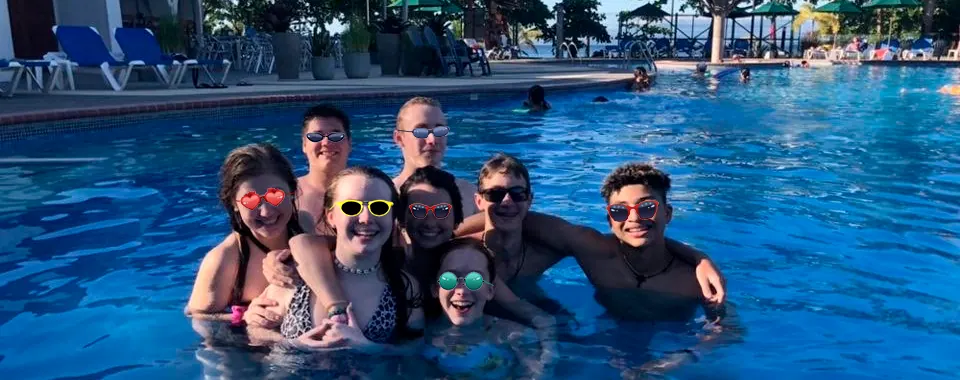 A group of friends in sunglasses grouped together in a resort pool, smiling at the camera.